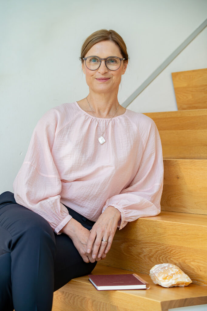 Arlett Hesse beim Personal-Branding-Shooting auf einer Holztreppe sitzend. Sie trägt eine rosafarbene Bluse, hat eine Brille auf und lächelt direkt in die Kamera. Entstanden während unseres Brandingshooting. 