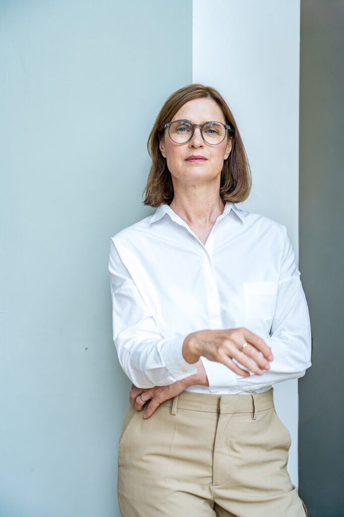 Arlett Hesse steht vor einer farblich weiß und mint abgesetzten Wand. Sie trägt eine weiße Bluse, eine Brille und die Haare offen. Sie schaut klar und ruhig geradeaus in die Kamera. Das Foto zeigt ihren Oberkörper. Entstanden während unseres Businessfotoshootings. 