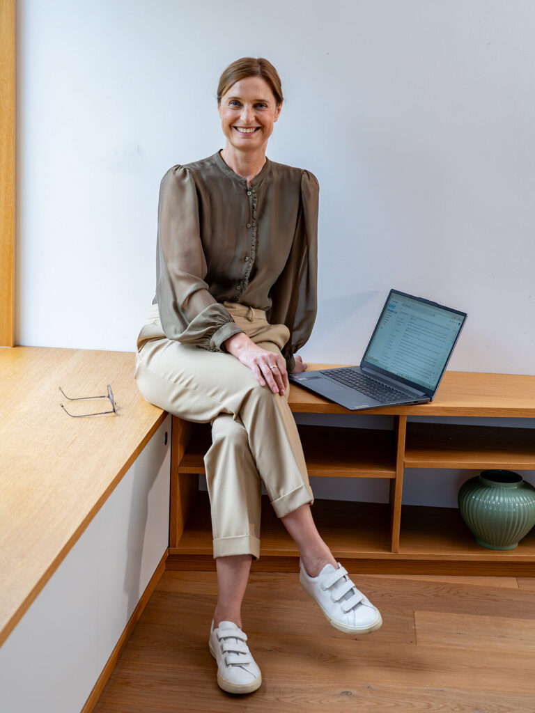 Arlett Hesse sitzt auf einem niedrigen Sideboard. Die Beine hat sie übereinandergeschlagen, ihre Brille liegt links im Bild neben ihr, der aufgeklappte Laptop steht rechts im Bild neben ihr. Die Haare hat sie zusammengebunden. Sie lächelt in die Kamera. Entstanden während unseres Fotoshootings.
