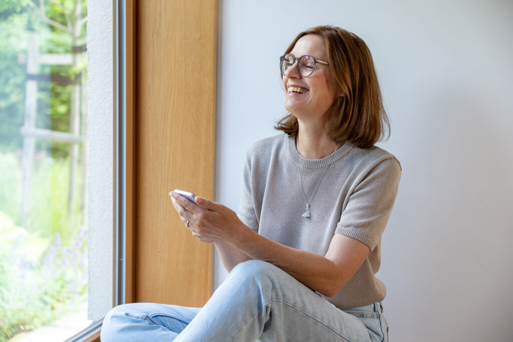 Arlett Hesse sitzend. Sie trägt eine helle Jeanshose und einen kurzärmeligen Pulli, hat eine Brille auf, trägt eine Brille und hält ein Handy in der Hand. Sie lacht herzlich während unseres Fotoshootings.